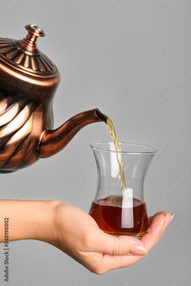 一名妇女将土耳其茶从茶壶倒进灰色背景的玻璃杯中