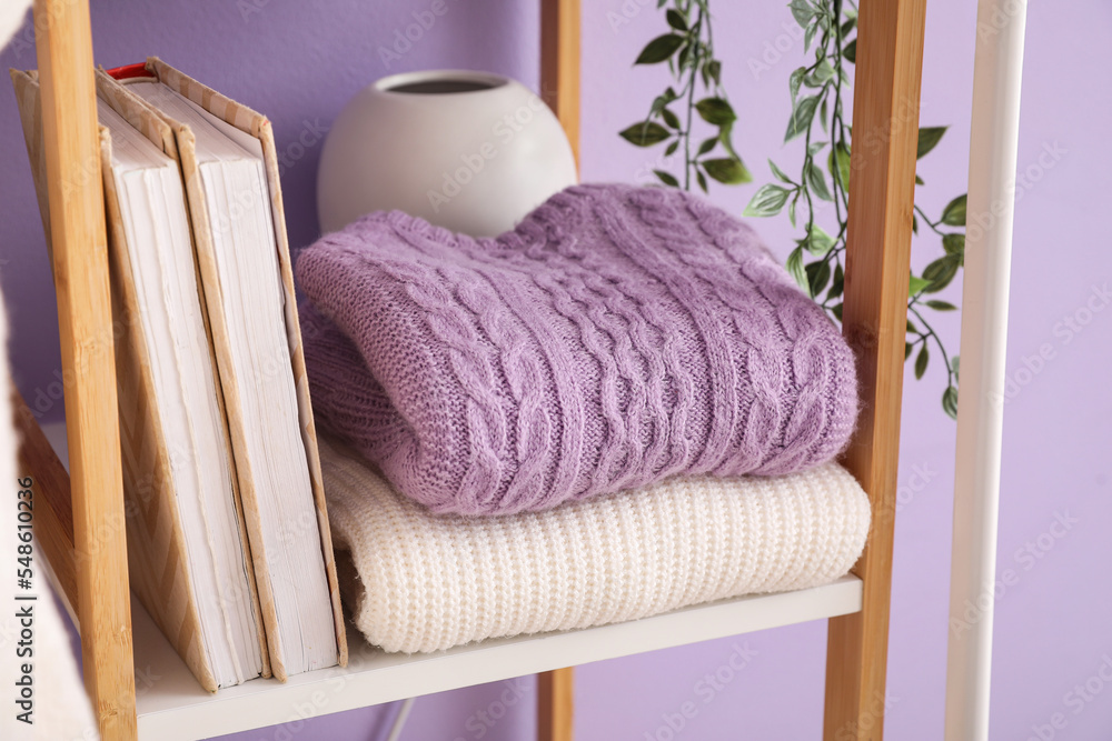 Knitted sweaters and books on shelf near lilac wall, closeup