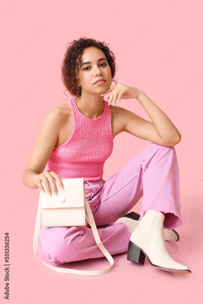 Beautiful African-American woman sitting and holding stylish handbag on color background