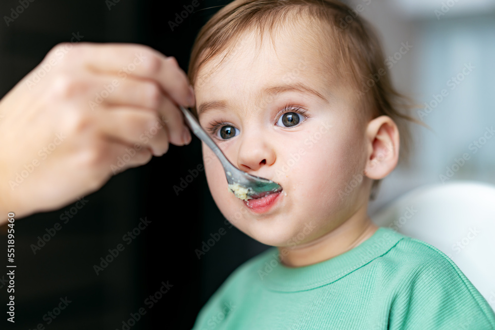 Mother feeding baby with spoon in kitchen. Small child sits on a chair and eating