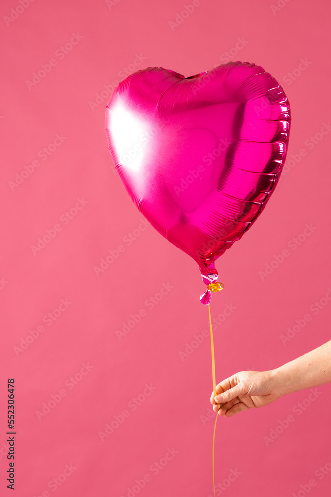Vertical of hand holding shiny pink heart shaped balloon on pink background with copy space