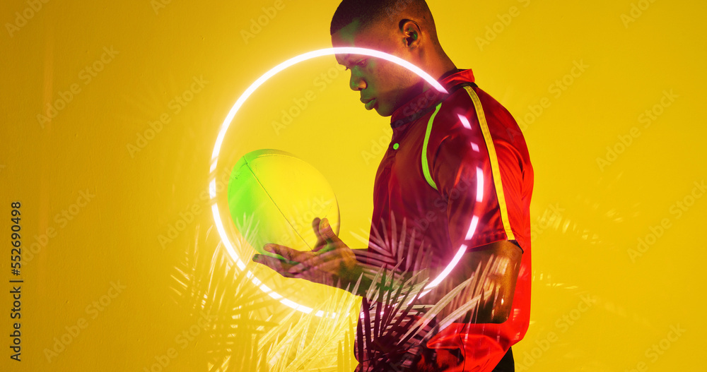 Side view of african american rugby player holding ball by circle and plants on yellow background