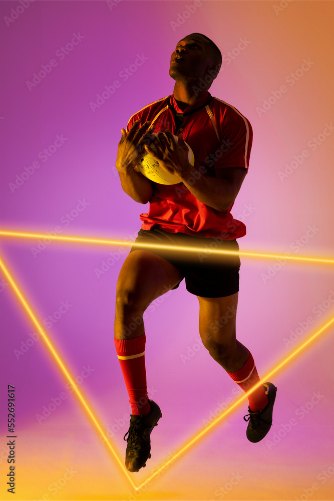 Full length of african american male rugby player catching ball and jumping by illuminated triangle