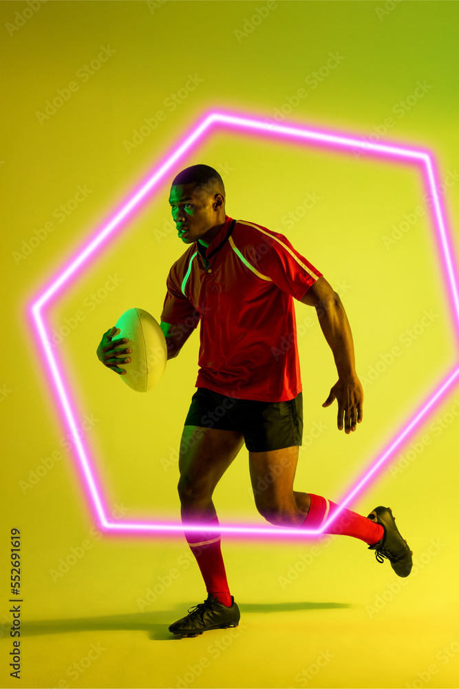 African american male rugby player with ball running by illuminated hexagon on green background