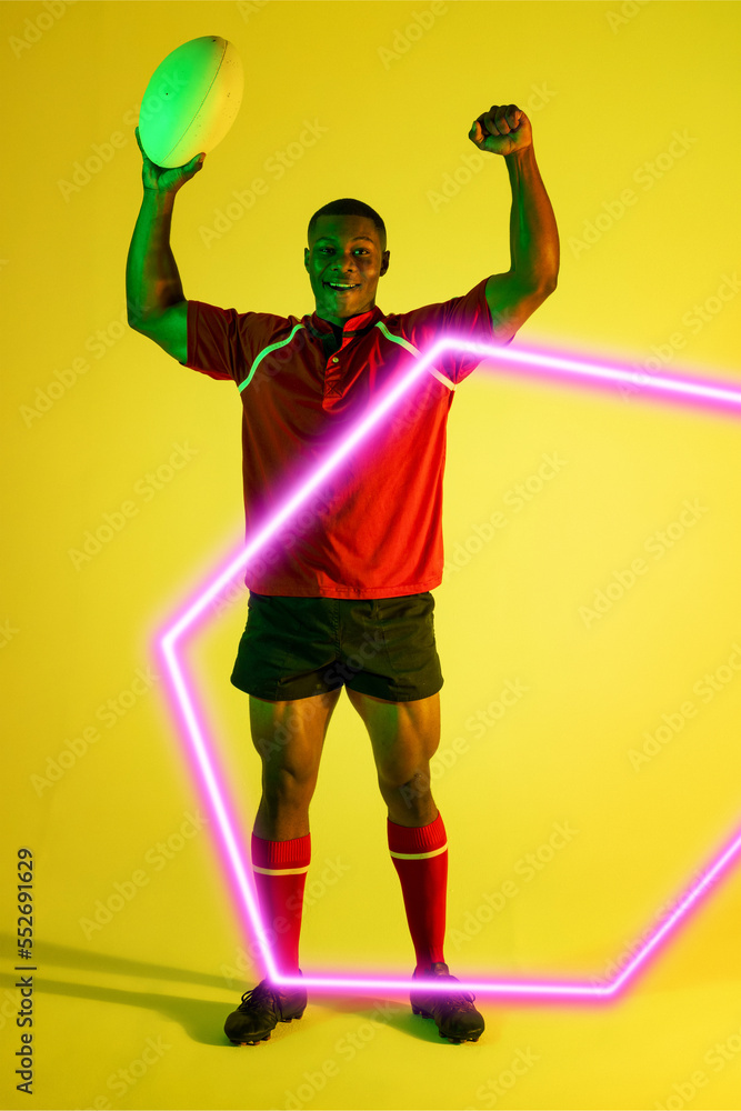 Smiling african american male player holding rugby ball and raising arms over illuminated hexagon