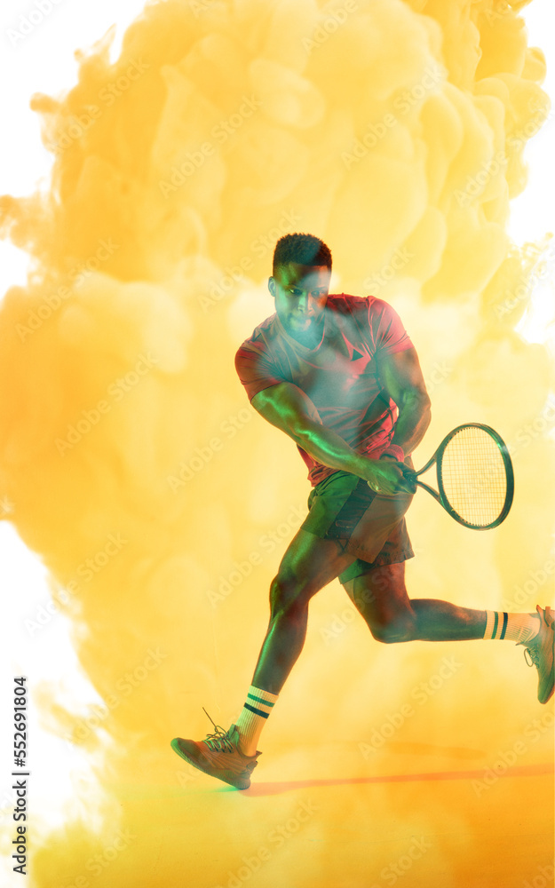 African american male tennis player running and hitting with racket against smoky yellow background