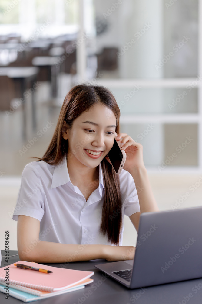 Beautiful Asian college student in uniform working with laptop computer and calling with smartphone.