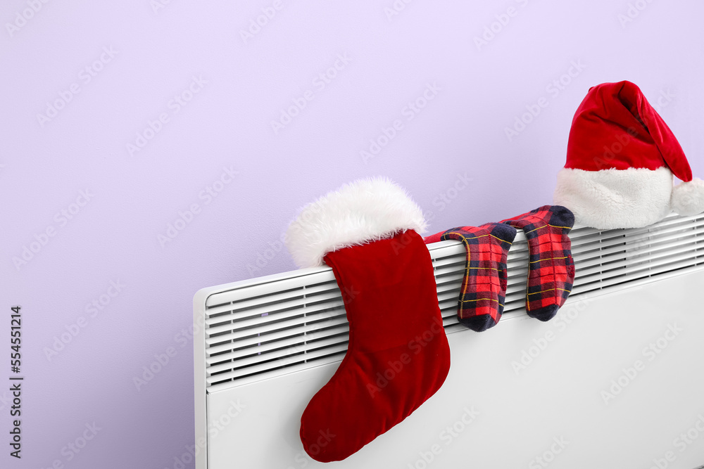 Santa hat and warm socks drying on electric radiator near lilac wall, closeup