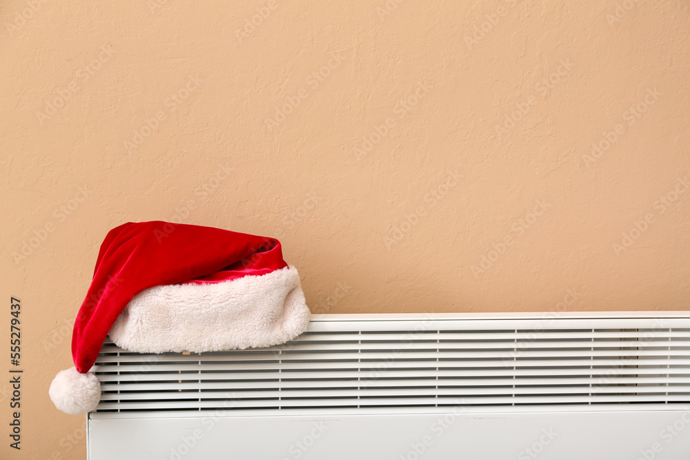 Santa hat on electric radiator near beige wall, closeup