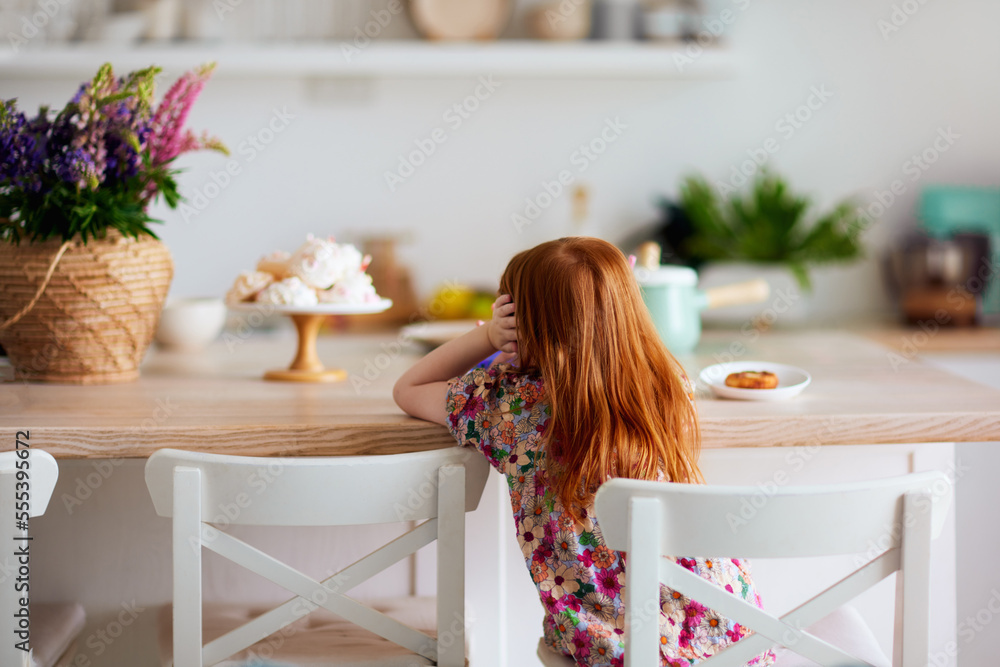 rear view of redhead baby girl sitting at the table on the kitchen at summer day