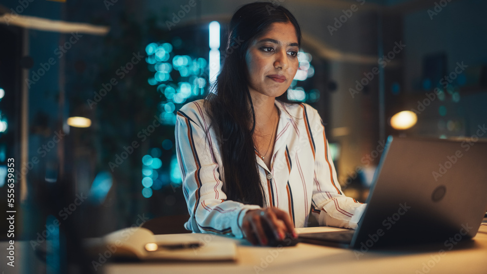 一位美丽的印度女性晚上在公司办公室用笔记本电脑工作的肖像。年轻