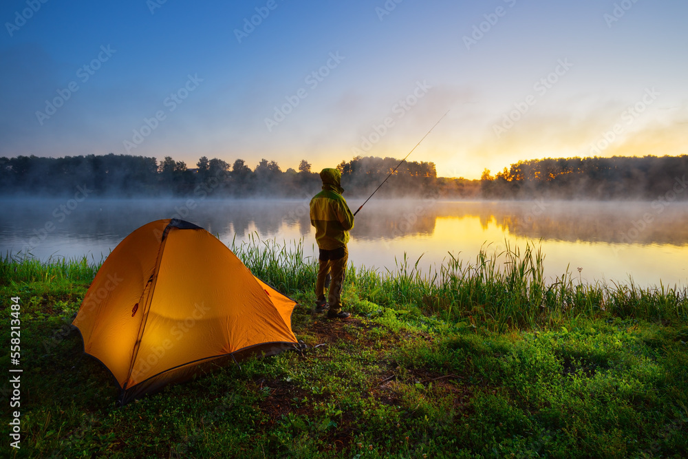 Fisherman on the bank of foggy river near an orange tent in the early morning