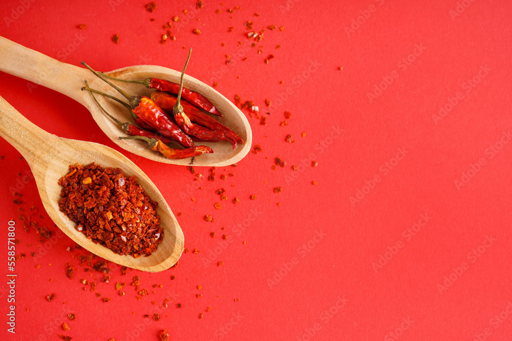 Spoons with chipotle chili flakes and dried peppers on red background