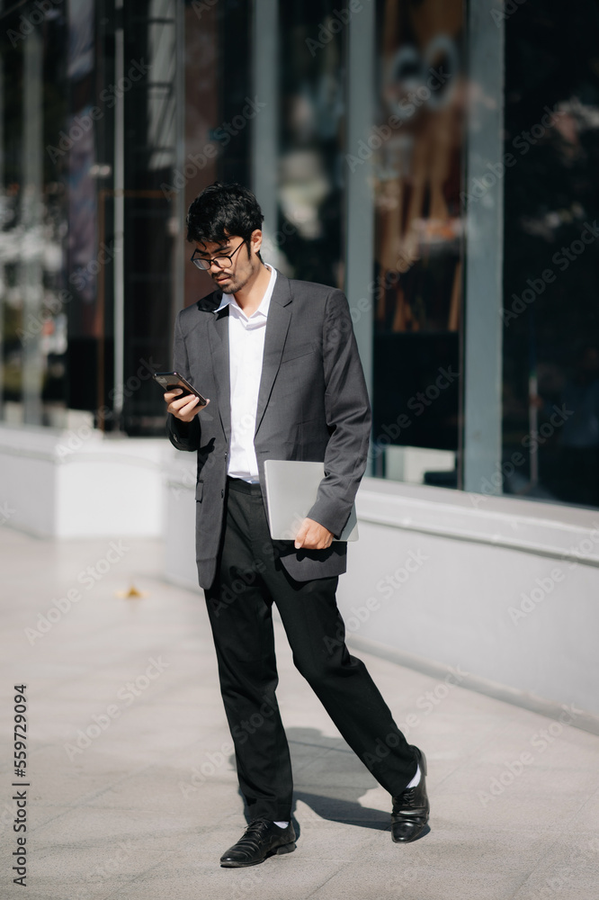 Handsome male entrepreneur standing outdoors with coffee cup and laptop in hands, leaving office in 