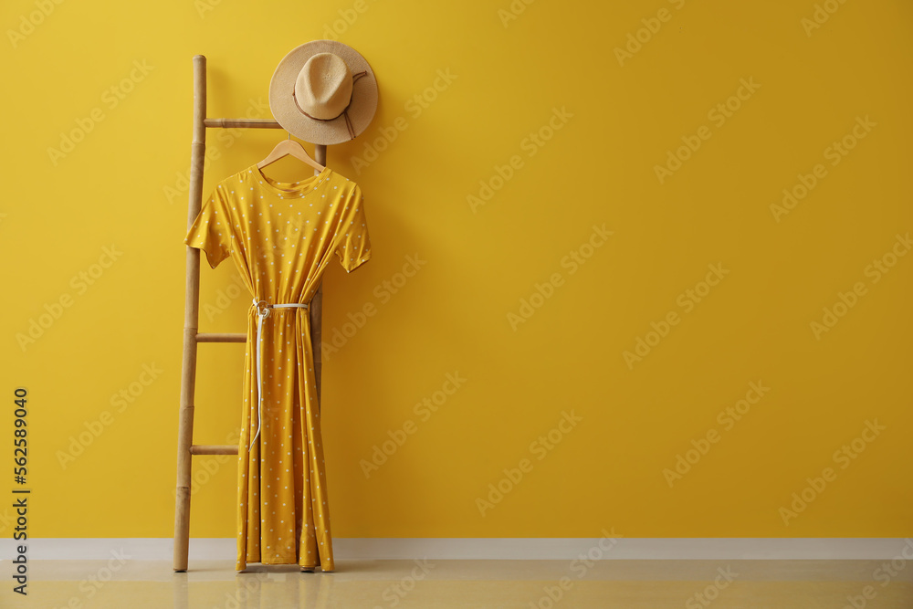 Ladder with stylish dress and hat near yellow wall