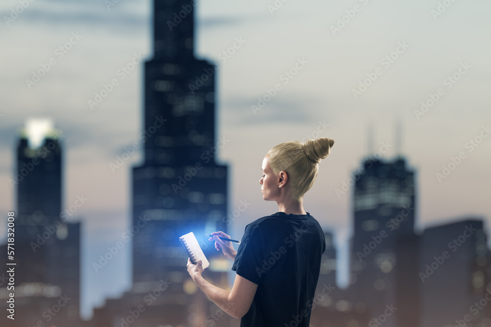 Attractive young european casual woman writing in glowing notepad on blurry city skyscrapers backgro