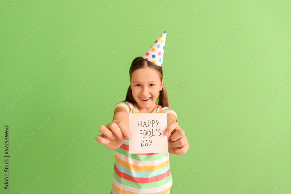 Little girl holding paper with text HAPPY FOOLS DAY on green background, closeup