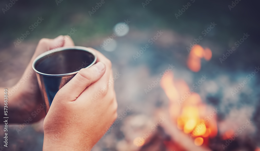 Humans hands holding the metal cup with hot drink above the fire.