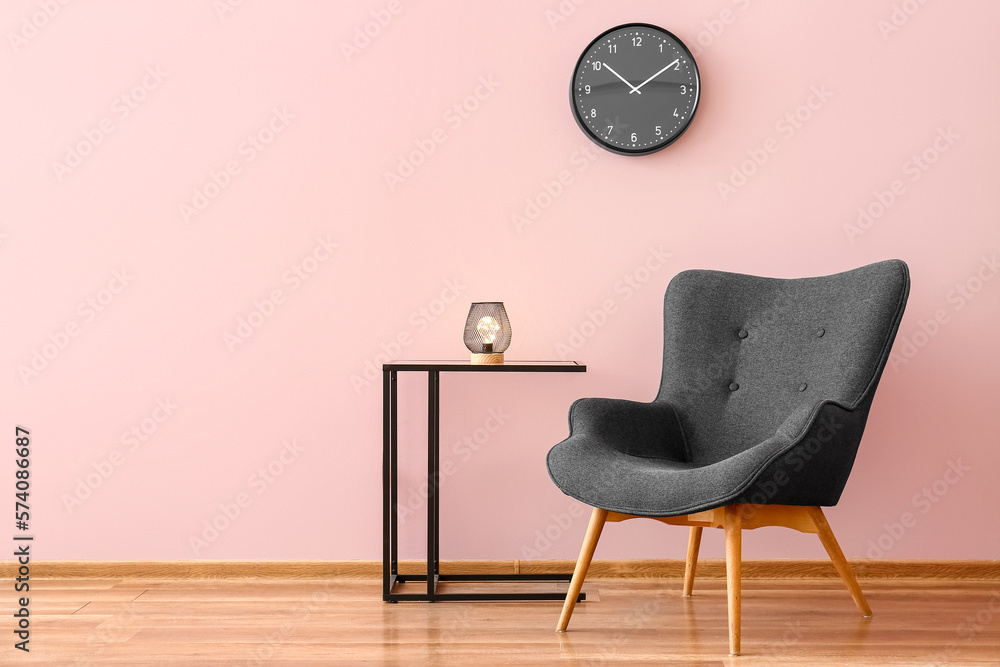 Stylish grey armchair, clock and glowing lamp on table near pink wall