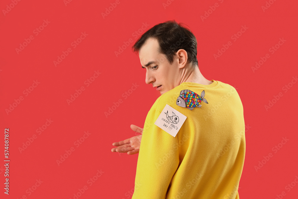 Sticky paper with text HAPPY FOOLS DAY and fish on young mans back against red background
