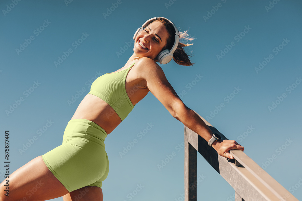 Fit and healthy woman smiling while listening to music with headphones and stretching her body outdo