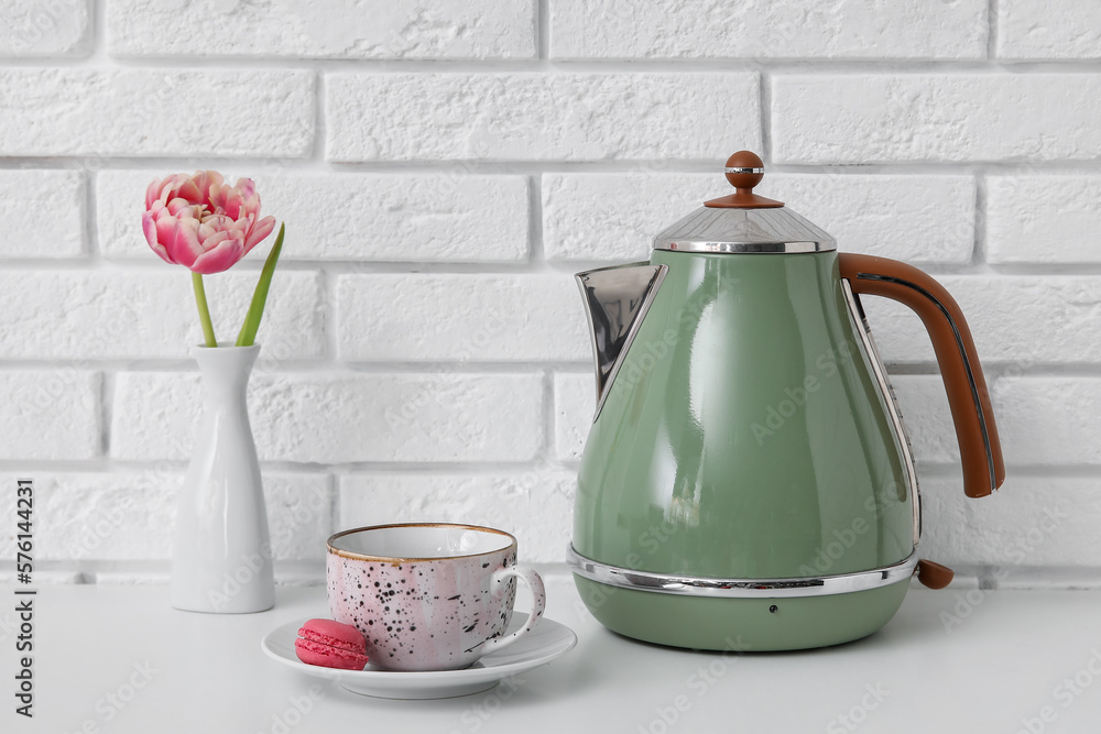 Electric kettle, cup, macaroon and vase with flower on table near white brick wall
