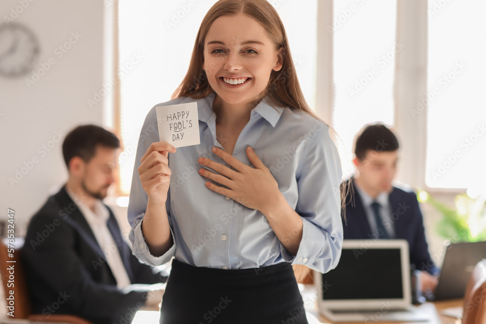 Young woman holding sticky paper with text HAPPY FOOLS DAY in office