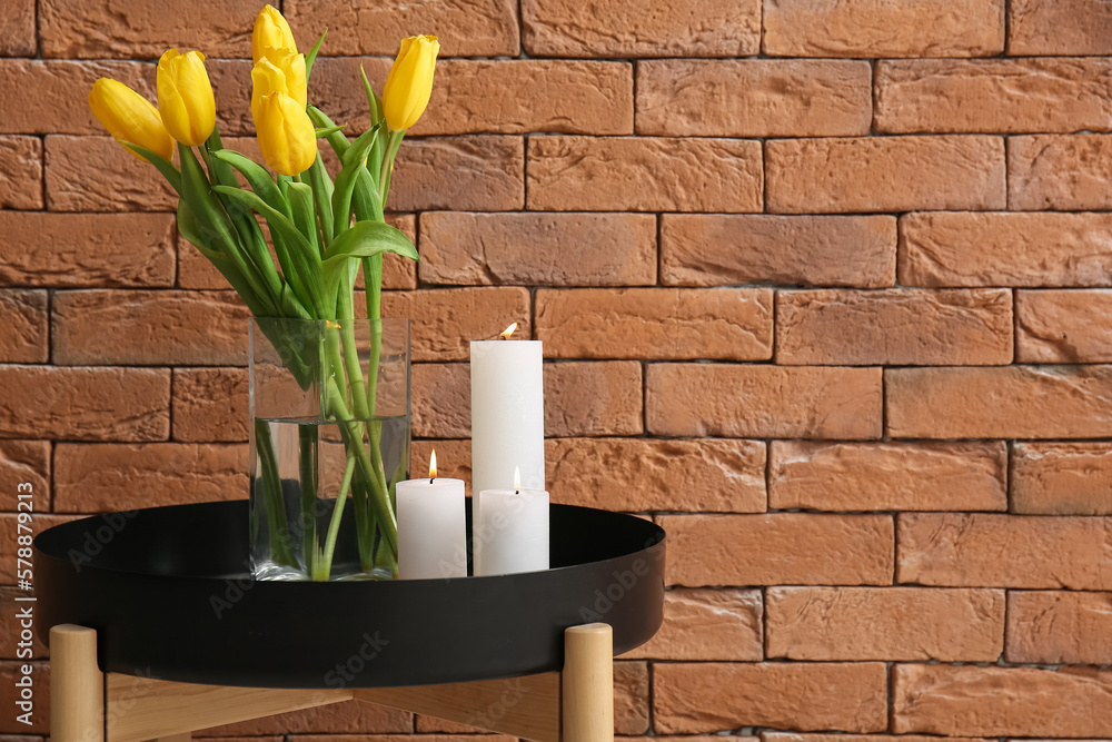 Burning candles and vase with tulip flowers on table near brown brick wall