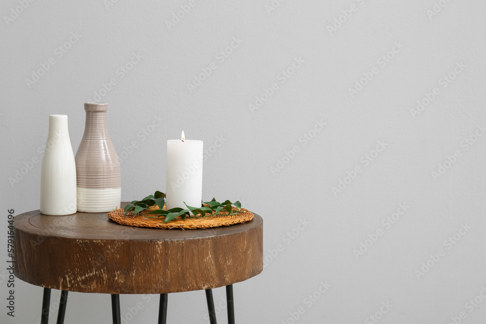 Burning candle, vases and ruscus branch on table near grey wall