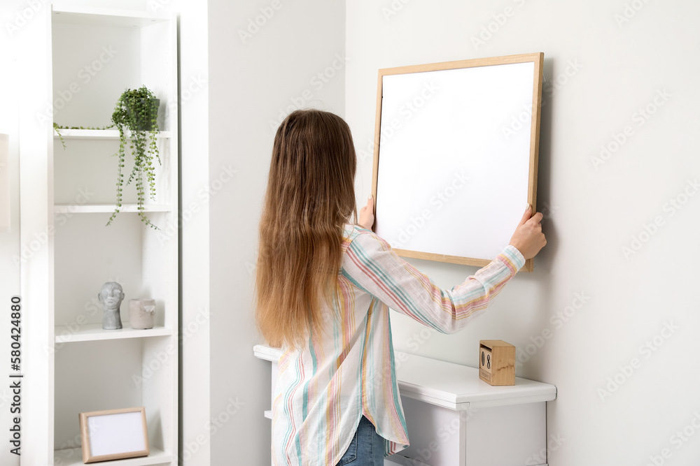 Young woman hanging blank frame on light wall at home