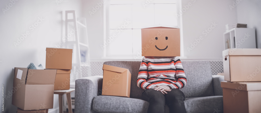 Tired happy woman with cardboard box on her head sitting on the sofa in her new house.