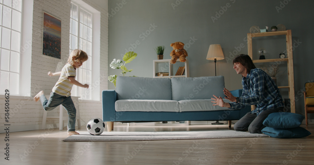 Funny father and son playing football at home. Little caucasian kid kicking the ball while his posit
