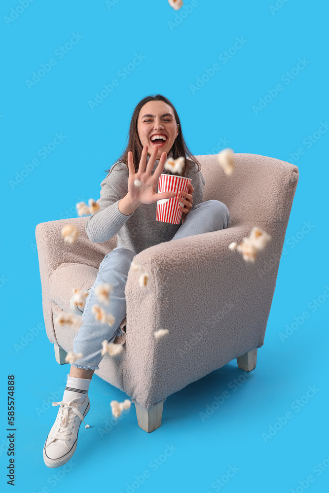 Young woman throwing popcorn in soft armchair on blue background