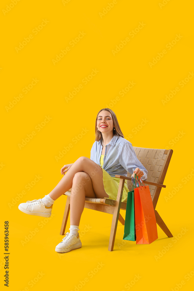 Young woman with shopping bags sitting in wooden armchair on yellow background