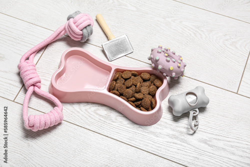 Set of pet care accessories and bowl of dry pet food on light wooden background