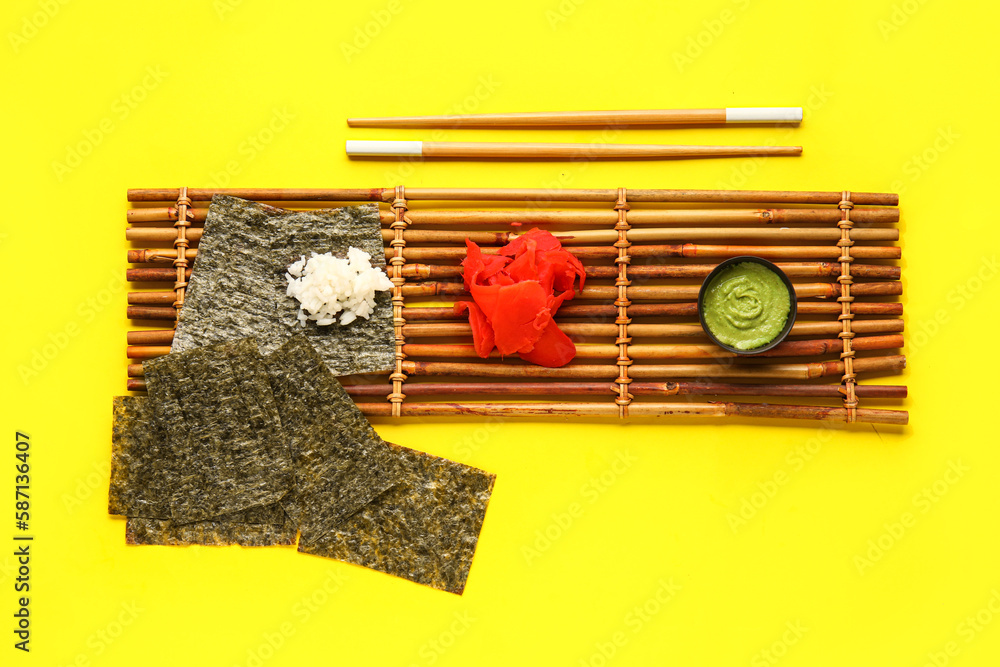 Bamboo mat with nori sheets, rice, ginger and wasabi on yellow background