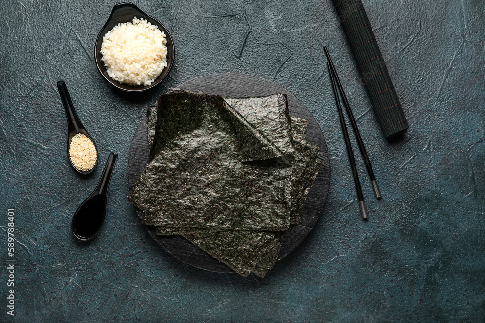 Board with nori sheets, rice, sesame seeds, soy sauce and sushi accessories on dark color background