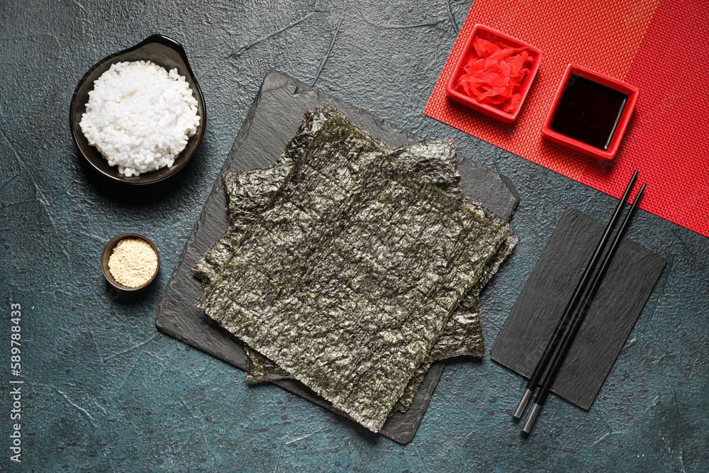 Board with nori sheets, rice, sesame seeds, ginger and soy sauce on dark color background