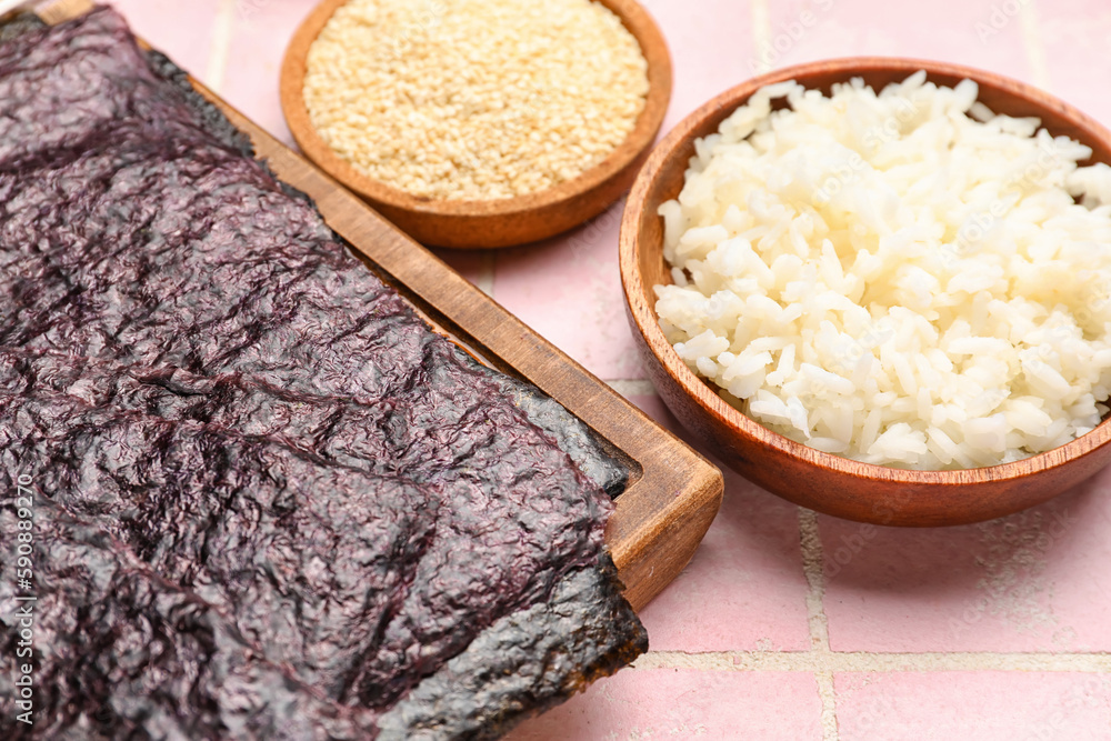 Wooden board with nori sheets and rice on pink tile background, closeup