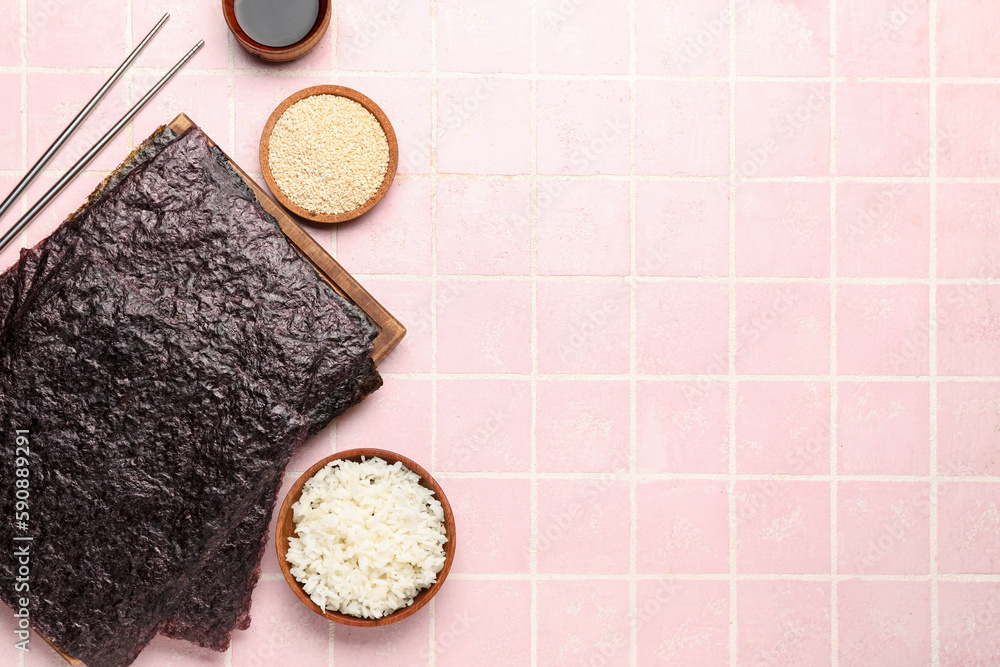 Wooden board with nori sheets, rice and sesame seeds on pink tile background