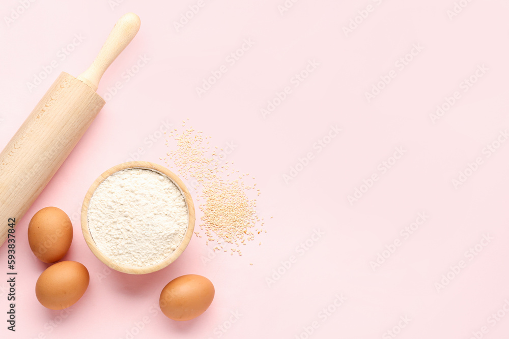 Bowl with wheat flour, sesame seeds, rolling pin and eggs on pink background