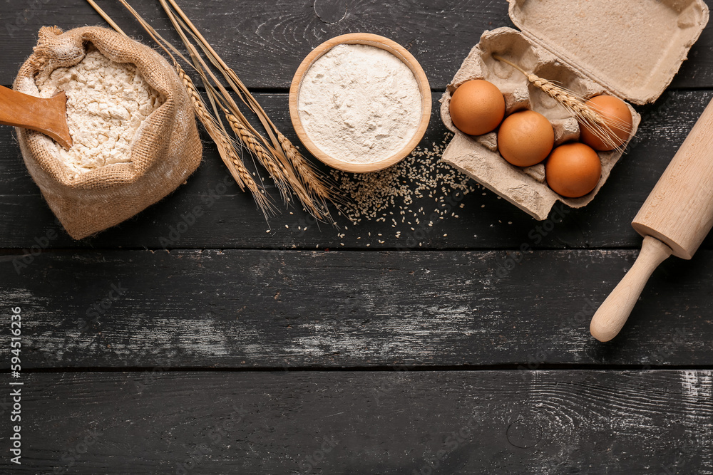 Composition with flour, wheat ears, eggs and rolling pin on black wooden table