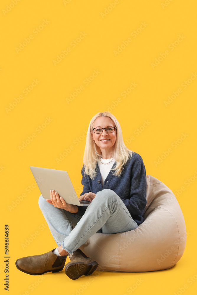 Mature female programmer working with laptop in beanbag on yellow background
