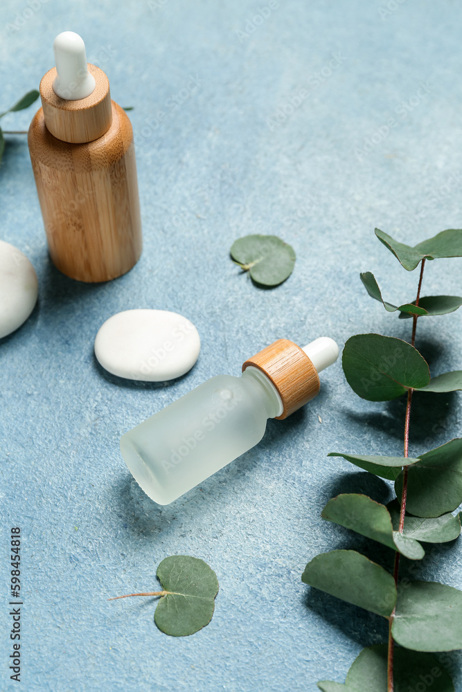 Bottles with cosmetic oil, eucalyptus branch and stones on blue textured background