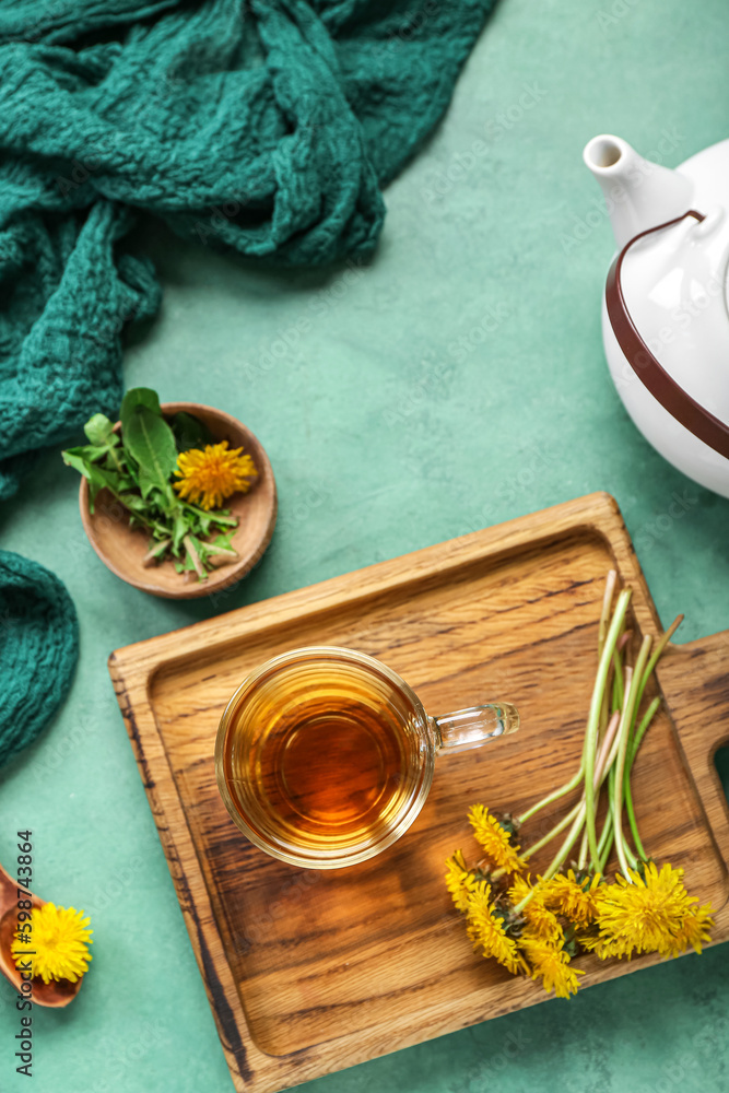 Board with glass cup of healthy dandelion tea and teapot on green background