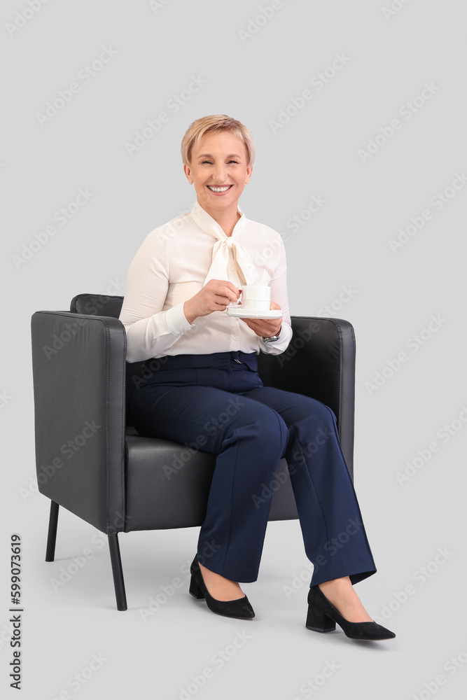 Mature businesswoman drinking coffee while sitting in armchair against grey background