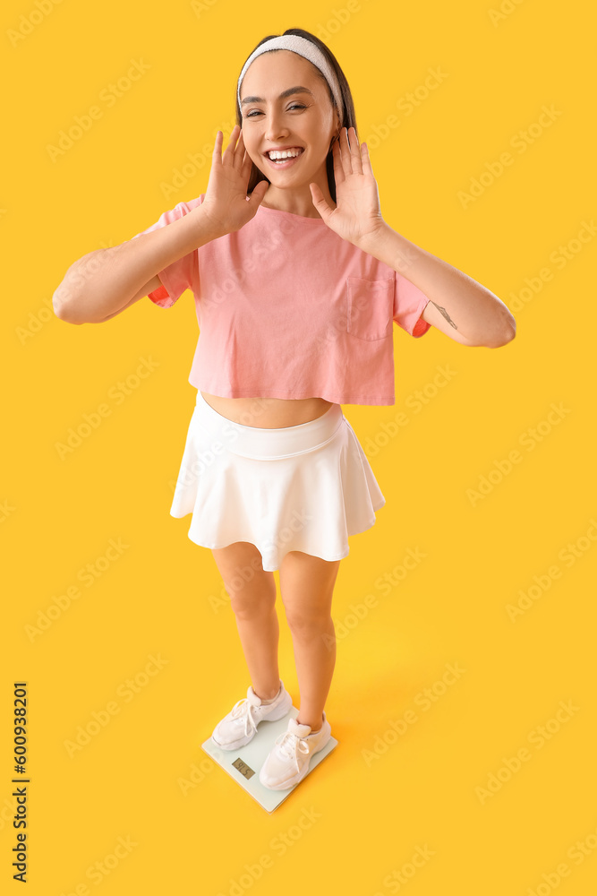 Young woman measuring her weight on scales against yellow background