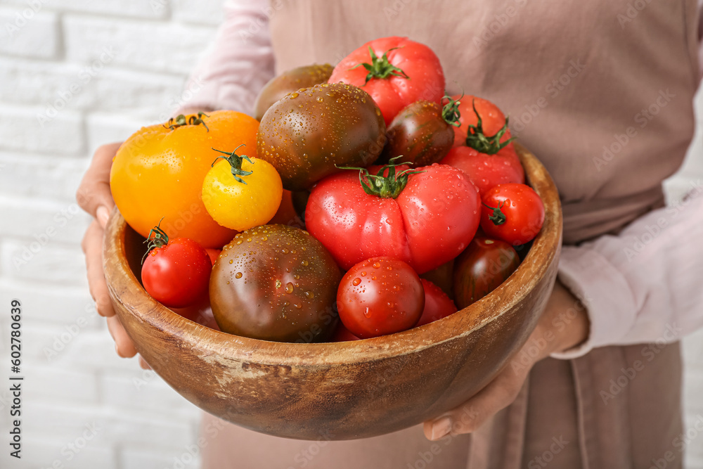 Woman holding wooden bowl with different fresh tomatoes on white brick background, closeup
