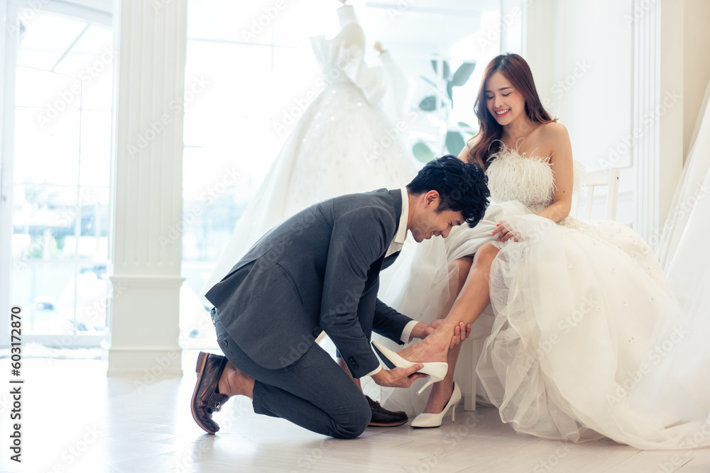 Asian beautiful bride trying on wedding shoes with her fiance in studio.
