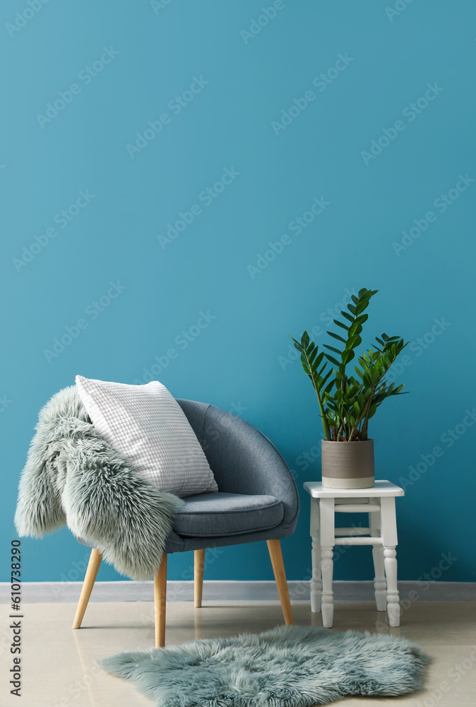 Cozy armchair with cushion, fur rug and houseplant on stool near blue wall